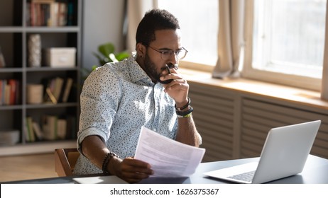 Focused Serious African American Businessman Accountant Analyst Holding Documents Looking At Laptop Computer Screen Doing Online Trade Market Tech Research Thinking Working Sit At Home Office Desk