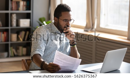 Focused serious african american businessman accountant analyst holding documents looking at laptop computer screen doing online trade market tech research thinking working sit at home office desk