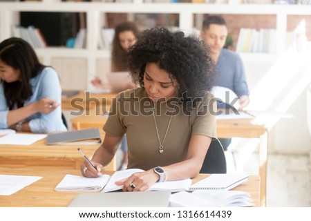 Focused serious african american female college high school student studying in multi ethnic classroom group writing essay in exercises book passing final exam test working on university assignment