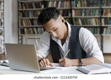 Focused Serious Asian Student Guy With Wireless Earphones Writing Notes, Research Article, Working On Study Project For Class In Library, Watching Online Learning Video, Webinar, Conference On Laptop