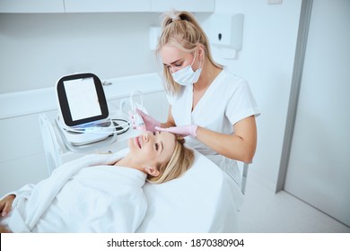 Focused serious Caucasian young female cosmetologist performing a beauty procedure using the fractional microneedle machine