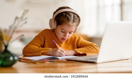 Focused serious elementary  little girl in headphones   making notes in notepad while communicating with teacher through video chat app during online education at home