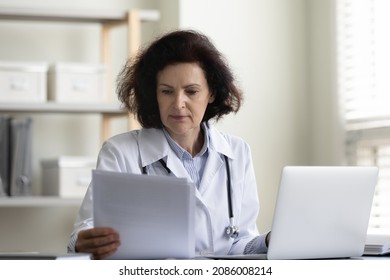 Focused serious middle aged GP doctor reading medical history, patient records, Chief physician reviewing insurance agreement, contract, paper documents at workplace with laptop. Medical job concept - Powered by Shutterstock