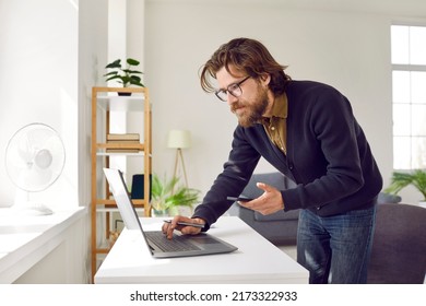 Focused Serious Smart Business Man With Laptop And Mobile Phone Working From Home. Bearded Man In Casual Clothes In Home Office Compares Information He Sees On Screen Of Laptop And Smartphone.