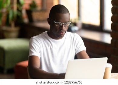 Focused Serious Young African Man Sitting Indoors Alone Working On Computer. Black American Millennial Smart Guy Wearing Glasses White T-shirt Looking On Notebook Screen Preparing For College Exams