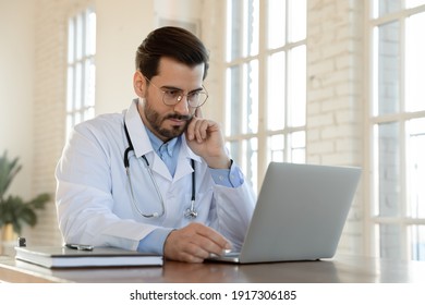 Focused Serious Young Male Doctor In Eyeglasses And White Uniform Working Online On Computer, Improving Medical Knowledge Or Giving Professional Consultation To Patient Distantly In Clinic Office.