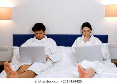 Focused serious young man and woman freelancers wearing in white bathrobes are working on laptops lying in bed in hotel room together. Freelance, distance, remote job concept, work and travel concept. - Powered by Shutterstock