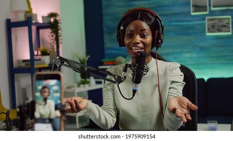 Focused shot on smartphone recording african blogger woman talking looking at camera during online podcast. Content creator streaming online broadcast, blogger discussing wearing headphones.