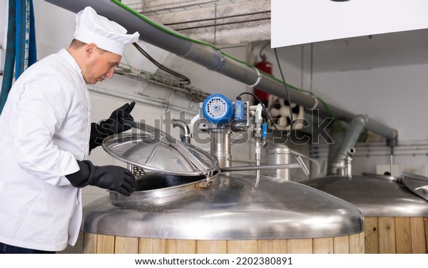 Focused skilled\
brewmaster opening lid of fermenter while checking process of craft\
beer production in\
workshop..