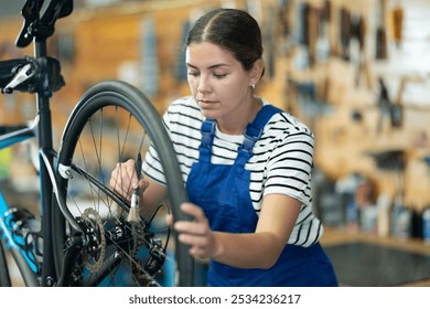 Focused skilled young female mechanic in blue overalls adjusting bicycle wheel, cleaning gears with small brush in professional bike repair shop - Powered by Shutterstock