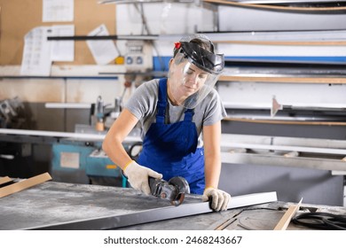 Focused skilled young woman in blue overalls, protective face shield and gloves using angle grinder to cut plastic profile in window manufacturing workshop - Powered by Shutterstock