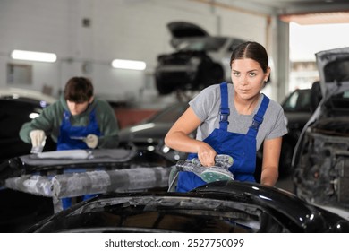 Focused skillful young female auto body technician, wearing blue overalls and a gray t-shirt, using hand sanding machine to smooth car bumper after puttying in workshop.. - Powered by Shutterstock