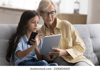 Focused smart granddaughter child helping grandma with online application on tablet, sitting on home couch, holding mobile phone, using digital gadgets for Internet communication - Powered by Shutterstock