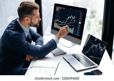 Focused smart successful caucasian male stock investor, broker, sits at his work desk, uses computer and laptop, concentrated analyzes risks and prospects, rise or fall of cryptocurrency coin