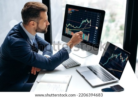 Focused smart successful caucasian male stock investor, broker, sits at his work desk, uses computer and laptop, concentrated analyzes risks and prospects, rise or fall of cryptocurrency coin