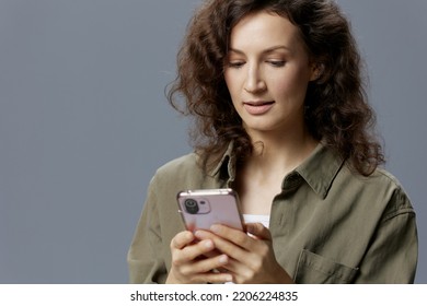 Focused Smiling Pensive Curly Beautiful Woman In Casual Khaki Green Shirt Typing New Post In Blog Using Phone Posing Isolated On Over Gray Blue Background. Social Media Influencer Concept. Copy Space