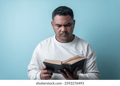 A focused Southeast Asian man in a white sweatshirt reads a book with a serious expression, deeply engaged in thought. Ideal for themes of learning, knowledge, and concentration.
