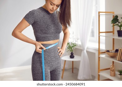 Focused sportswoman checks her waist size with measuring tape, emphasizing importance of fitness. Young woman in stylish tracksuit measures her waist with measuring tape in her bright living room. - Powered by Shutterstock