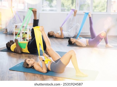 Focused sporty young girl performing set of exercises with resistance band during pilates class with mixed age group of women in light airy fitness studio - Powered by Shutterstock