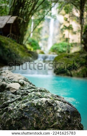 Focused stone in foreground as product stage with blurred jungle in background. In the background is a pond with turquoise water, waterfall and trees.