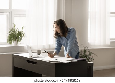 Focused student woman stands at desk do homework, prepare for exams, check information, take notes, writes thesis. Young freelancer working on financial report, analyzes statistical data using laptop - Powered by Shutterstock