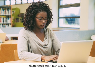 Focused student working in library computer class. Young black woman sitting at desk and using laptop in classroom with bookshelves. Education concept