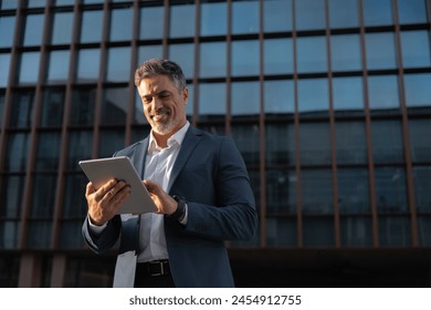 Focused successful mature Indian or Latin entrepreneur businessman holding digital pc tablet standing outdoor at business office building. Hispanic smiling man in suit working using touchpad computer - Powered by Shutterstock