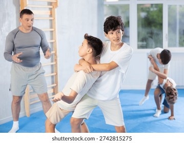 Focused teen boy practicing basic protection skills with opponent in training bout during self defense course with coach in gym, trying to escape rear bear hug technique..