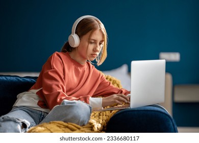 Focused teen girl wearing wireless headphones sitting on sofa with laptop, studying online, having virtual lesson. Child teenager surfing internet, learning something new  - Powered by Shutterstock