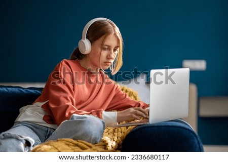Focused teen girl wearing wireless headphones sitting on sofa with laptop, studying online, having virtual lesson. Child teenager surfing internet, learning something new 