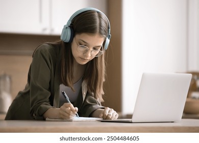 Focused teenage girl prepare for exam at home, standing at table in kitchen, listen audio course on wireless headphones, writes notes, learn foreign language using on-line lesson, studying on internet - Powered by Shutterstock