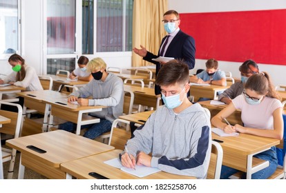 Focused Teenage Students In Protective Face Masks Studying In Classroom With Teacher, Writing Lectures In Workbooks. Necessary Precautions In Coronavirus Pandemic