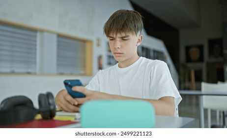 A focused teenager boy using a smartphone while sitting at a table in a campus library - Powered by Shutterstock