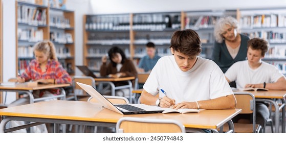 Focused teenager male student sitting at library desk with laptop, preparing for exam - Powered by Shutterstock