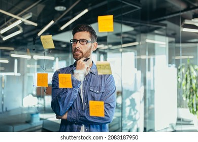 focused and thinking businessman writing on colorful notes attached to a glass wall, the entrepreneur creates a visual to-do list, business goals for future careers, daily goals