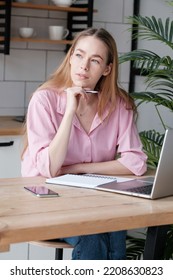 Focused Thoughtful Attractive Young Woman Sits At Table In Kitchen In Front Of Notepad, Smartphone And Laptop And Looks Away. Freelance Employee Working At Computer, Thinking Over Ideas