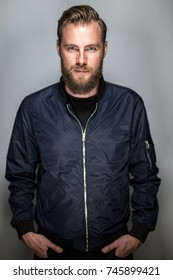 Focused Tough Man In Beard And Blue Jacket, Standing Against A Grey Background Looking At Camera. Short Depth Of Field.