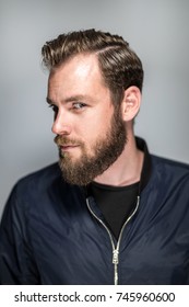 Focused Tough Man In Beard And Blue Jacket, Standing Against A Grey Background Looking At Camera. Short Depth Of Field.