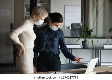 Focused two young multiracial business women in medical facial protective masks standing near table, looking at laptop screen, discussing project details, preventing spreading corona virus in office. - Powered by Shutterstock