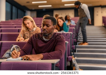 Focused university student writing notes during a lesson in a bustling lecture hall, fully engaged in the learning experience