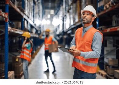 A focused warehouse supervisor observes the inventory with a digital tablet, wearing a safety helmet and vest. His thoughtful expression highlights his role in inventory management. - Powered by Shutterstock