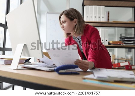 Focused woman compares paper accounting documents with computer version standing at table with big monitor. Female accountant does paperwork in light office