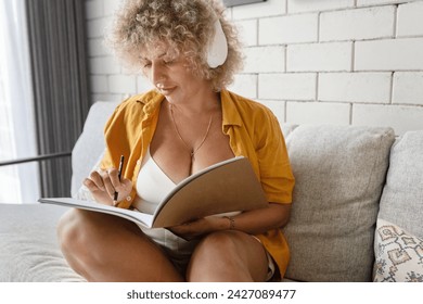 A focused woman with curly hair and headphones jotting down notes in a notebook while sitting comfortably on a couch at home. - Powered by Shutterstock