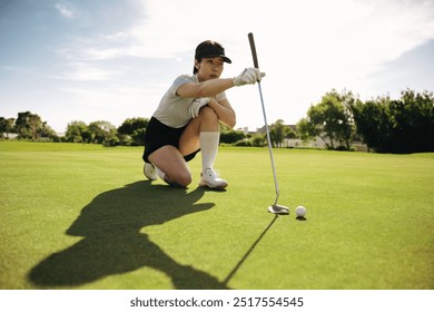 Focused woman golfer kneeling and lining up a putt on a bright, sunny day outdoors at a lush golf course. - Powered by Shutterstock