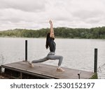 Focused woman practicing yoga on dock by serene lake, demonstrating warrior pose amidst nature Health and wellness concept with tranquility and strength