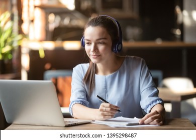 Focused woman wearing headphones using laptop in cafe, writing notes, attractive female student learning language, watching online webinar, listening audio course, e-learning education concept - Shutterstock ID 1395298487