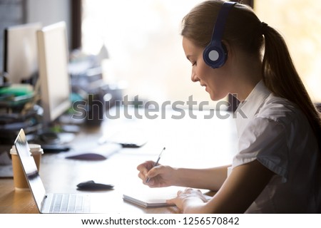 Focused woman wearing headphones write notes watch webinar study online with online teacher, young female student learning computer course with laptop listening lecture, interpreter translating class
