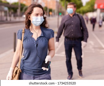 Focused Woman Wearing Medical Mask And Rubber Gloves Listening To Music On Earphones On Way To Work Along City Street On Spring Day. New Life Reality During COVID 19 Pandemic