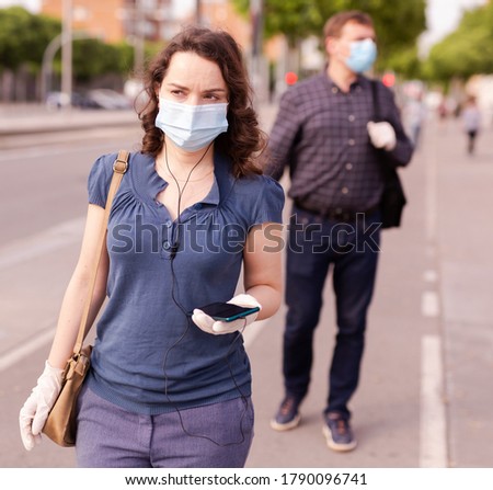 Focused woman wearing medical mask and rubber gloves listening to music on earphones on way to work along city street on spring day. New life reality during COVID 19 pandemic