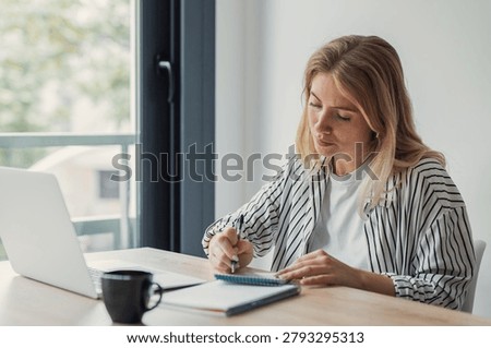 A focused woman writes in a notebook while working on her laptop at a bright home office desk with coffee and documents, conveying productivity, remote work, concentration and study.